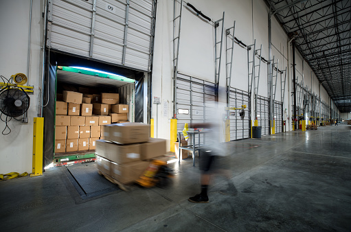 Long exposure view of warehouse workers using pallet jacks to unload a truck at a loading dock in a fulfillment center.