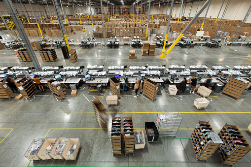 High angle view of lines of packing stations in a fulfillment center.  A team of packers are packaging up orders brought to them on carts from the picking area, then throwing them onto a conveyor belt.