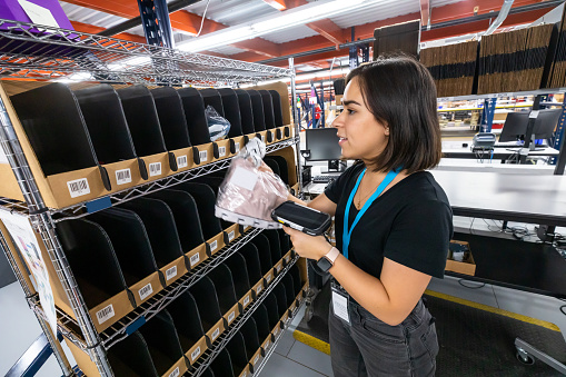 A young woman working as a packer in a fulfillment center, taking items of clothing out of a picking cart and scanning them with a barcode scanner.