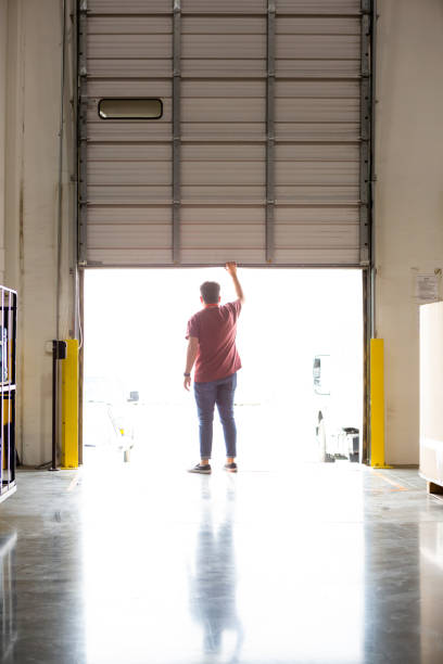 Fulfillment Center Worker Opening Truck Loading Dock Rear view of a warehouse employee pushing open the door of a loading dock in preparation for a truck arriving, letting bright sunlight inside. loading bay stock pictures, royalty-free photos & images