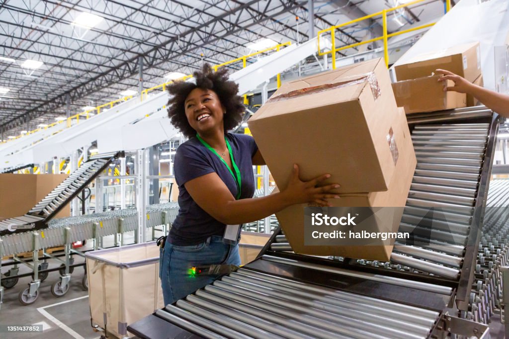 Cheerful Warehouse Employee Loading Boxes Into Truck A grinning woman collecting boxes at the bottom of a sorting chute and pushing them along an extendable conveyor belt towards a truck loading dock for delivery. Freight Transportation Stock Photo