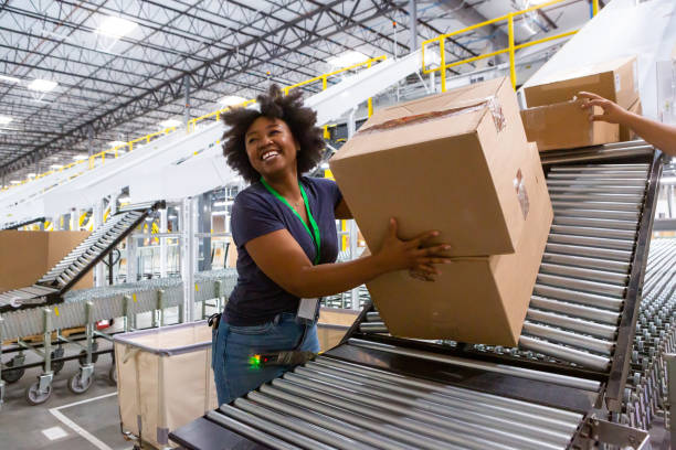 Cheerful Warehouse Employee Loading Boxes Into Truck A grinning woman collecting boxes at the bottom of a sorting chute and pushing them along an extendable conveyor belt towards a truck loading dock for delivery. freight transportation stock pictures, royalty-free photos & images
