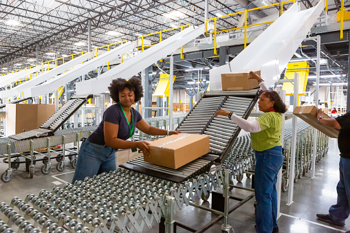 Fulfillment center workers pushing packages along an extendable conveyor belt from the bottom or a sorting chute into a truck.
