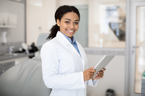 Modern Dentistry. Portrait Of Smiling Black Dentist Female Doctor With Digital Tablet In Hands Standing At Workplace In Modern Clinic Interior, Professional Stomatologist Lady Looking At Camera