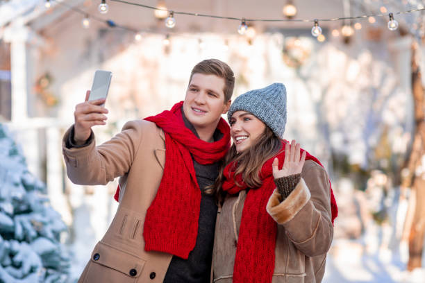 Beautiful lovers happy man and woman celebrating winter holidays together, standing at snowy backyard, hugging and holding mobile phone, having video chat with friends, copy space