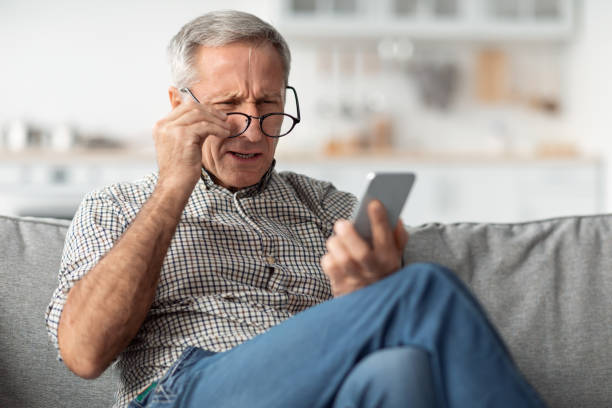 hombre mayor entrecerrando los ojos leyendo mensaje usando anteojos en casa - entrecerrar los ojos fotografías e imágenes de stock