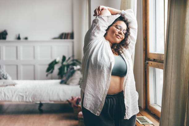 Curly haired overweight young woman in blue top and shorts with satisfaction on face accepts curvy body shape in stylish bedroom. stock photo
