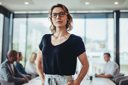 Portrait of a confident business woman standing in office with coworkers talking in the background in meeting room.