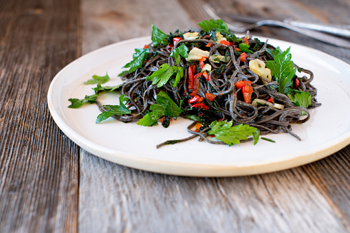 Gluten free and vegetarian, spaghetti dish cooked with black bean pasta, sliced garlic, olive oil, bell peppers and parsley. Served isolated on a plate on wooden table. Closeup