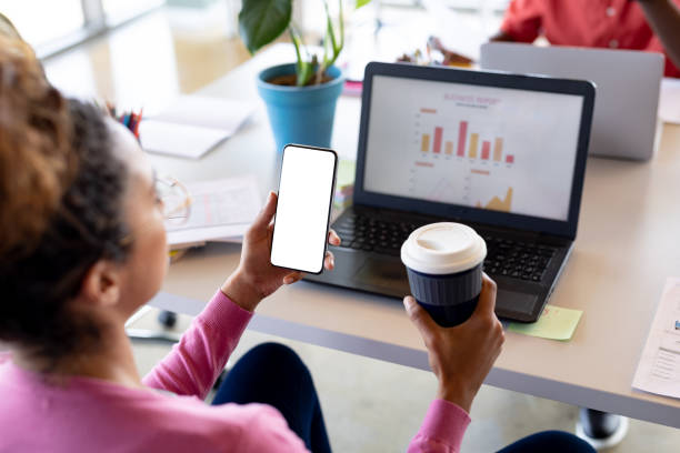 istock Businesswoman holding coffee cup while looking at smartphone with copy space in office 1351448179