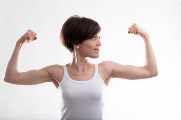 Pretty young woman wearing a sleeveless top while flexing her arms for showing her biceps muscles as a gesture of power and confidence against white background for copy space
