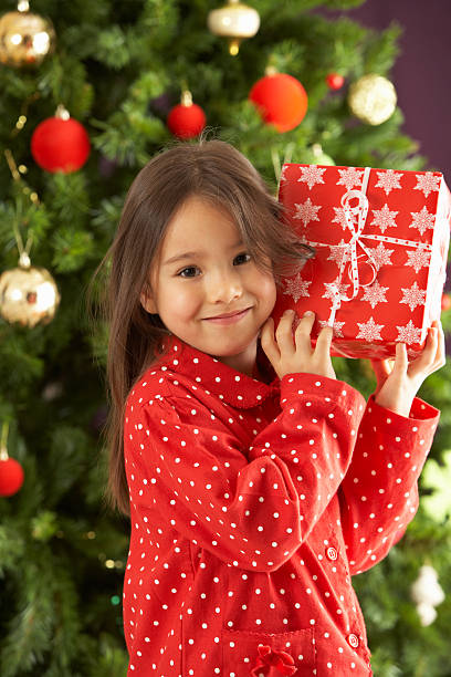 istock Young Girl Holding Wrapped Present In Front Of Christmas Tree 135145167