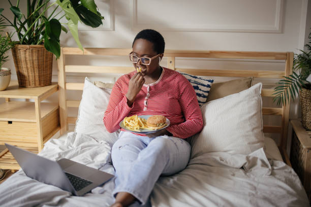 istock Young woman working or studying from home while sitting comfortably on her bed; Eating fast food, hamburger and french fries 1351455513