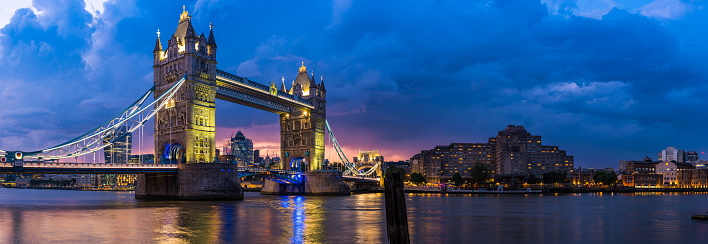 The iconic gothic battlements of Tower Bridge framing the modern skyscraper skyline of the City of London illuminated against a dramatic sunset sky over the River Thames in the heart of the UK’s vibrant capital city.