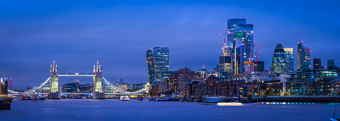 The futuristic spires of the City Financial District skyscrapers overlooking the iconic battlements of Tower Bridge and the River Thames illuminated at night in the heart of London, UK.