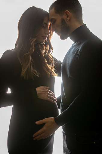 Beautiful couple posing in studio. She is pregnant in last trimester and looks amazing. They both wear black clothes. Strong backlight