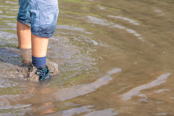 istock Young boy with short blue trowsers wading with wet socks and wet boots through high tide after a floodwater has broken the dike and overflown the lands behind 1351458178