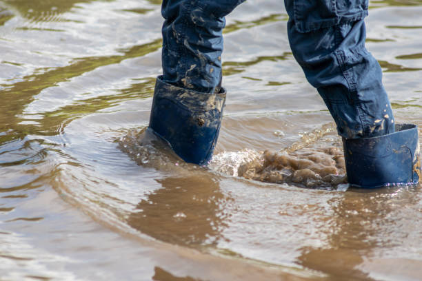 kleiner junge mit kurzen blauen trowsern, die mit nassen socken und nassen stiefeln durch flut waten, nachdem ein hochwasser den deich durchbrochen und das land dahinter überfloben hat - überschwemmung stock-fotos und bilder