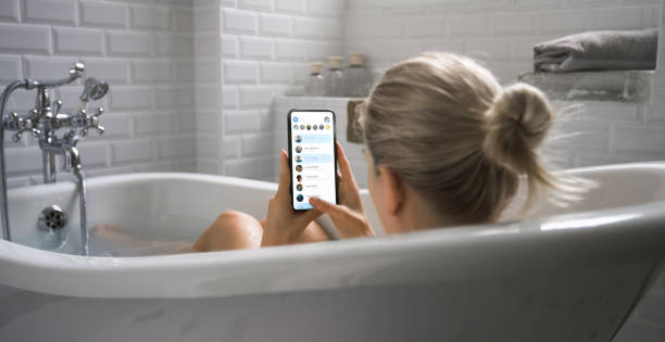 bella ragazza bionda fa il bagno usa lo smartphone per controllare i social network mentre si rilassa. la ragazza attraente utilizza il telefono cellulare per navigare sui social media, chattare con gli amici, navigare in internet. - sensuality people mobile phone 30s foto e immagini stock