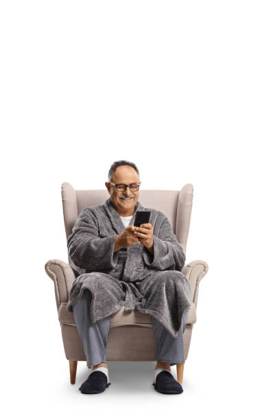 SMiling mature man in a bathrobe sitting in an armchair and typing on a smartphone SMiling mature man in a bathrobe sitting in an armchair and typing on a smartphone isolated on white background man sleeping chair stock pictures, royalty-free photos & images