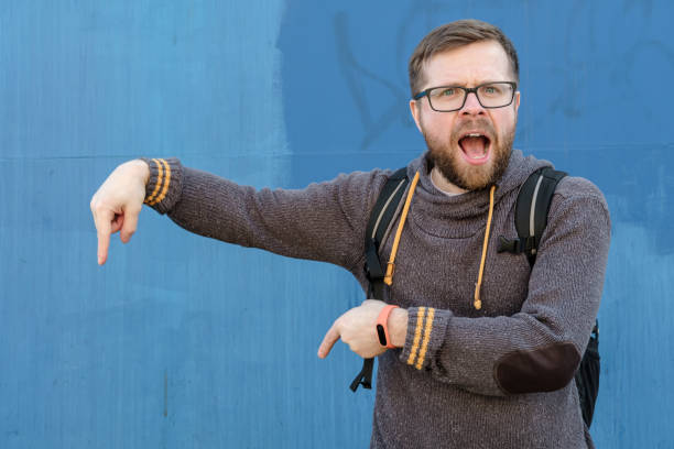 istock Man in casual clothes with a city backpack points index finger down and speaks out loud while looking at the camera. Copy space. 1351462077
