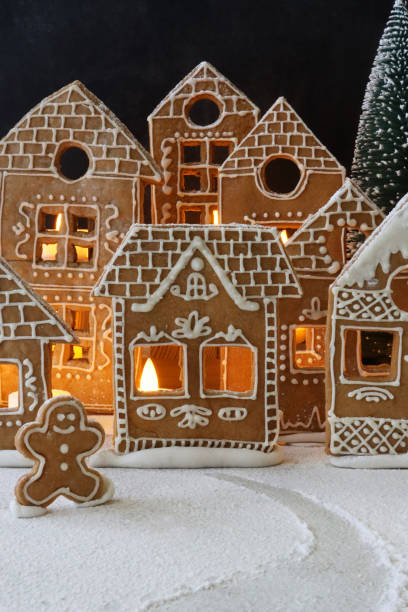 istock Image of homemade, gingerbread house biscuits decorated with white royal icing displayed in snowy, night Christmas village scene, illuminated by fairy lights, gingerbread man cookie, icing sugar snow, black background 1351467455