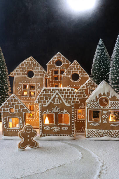 istock Image of homemade, gingerbread house biscuits decorated with white royal icing displayed in snowy, night Christmas village scene, illuminated by fairy lights, gingerbread man cookie, icing sugar snow, black background with full moon 1351467477