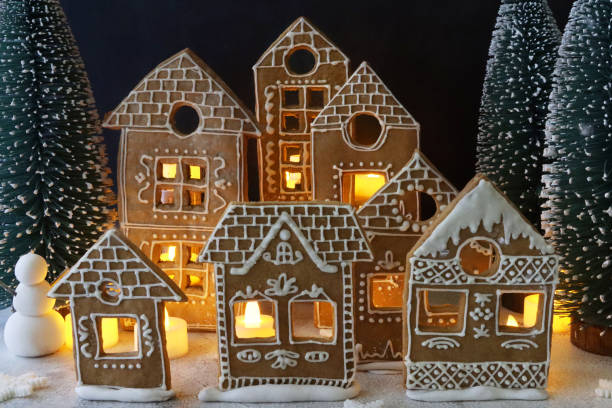 istock Image of homemade, gingerbread house biscuits decorated with white royal icing displayed in snowy, night Christmas village scene, illuminated by fairy lights, snowman, icing sugar snow, black background 1351467478