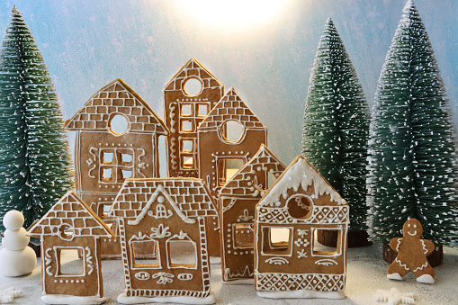 Stock photo showing close-up view of a snowy night-time, Christmas village scene. Homemade, gingerbread house cookies decorated with white royal icing, illuminated with fairy lights and surrounded by model fir trees on white, icing sugar snow against a blue snowy background.