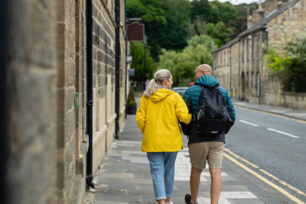 Bonding and Exploring Rearview of a couple wearing raincoats walking around and exploring Warkworth, Northumberland. They are looking at each other while talking. arm in arm stock pictures, royalty-free photos & images