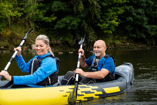 A couple wearing activewear and life jackets kayaking in a river in Warkworth, Northumberland. They are using oars to paddle along the river and they are smiling.