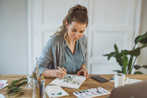 Mature woman at home painting. She is watching tutorial on computer and painting. Sitting on kitchen counter in front of window.