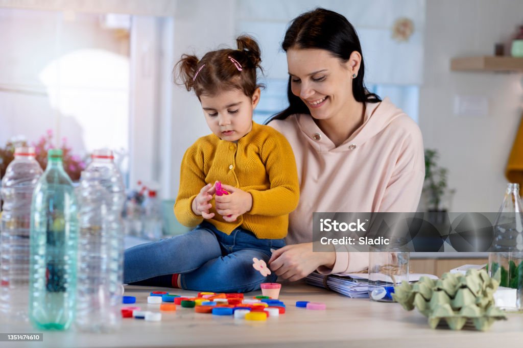 Have a fun and get the job done! Mother teaching her daughter how to recycle waste. Beautiful woman education her cute little daughter  and playing  with her. Sorting garbage at home. Bottle Cap Stock Photo