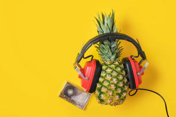 Photo of vinyl record, magnetic audio tape and pineapple in red retro headphones on yellow background, music lover concept