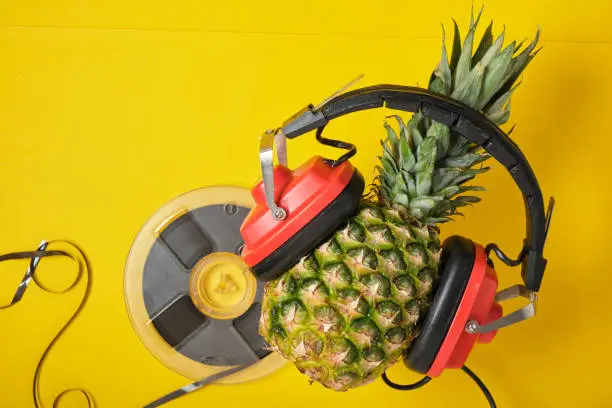 Photo of magnetic audio tape reel and pineapple in red retro headphones on yellow background, music lover concept