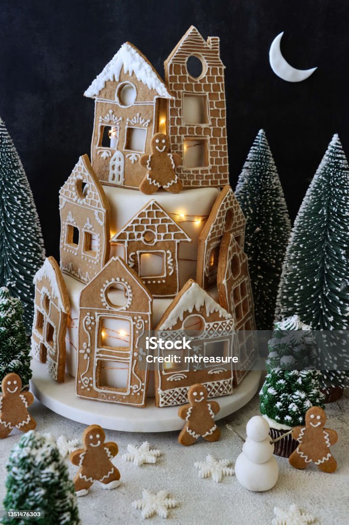 Image of homemade, tiered Christmas cake displayed on cake stand, cake covered with fondant icing and gingerbread house biscuits decorated with white royal icing, illuminated fairy lights, night-time Christmas village scene, black background and crescent Stock photo showing close-up view of a night-time Christmas village scene designed Christmas cake. Homemade, tiered cake covered in fondant icing and gingerbread house-shaped cookies decorated with white royal icing displayed on cake stand against a black background. Christmas Stock Photo