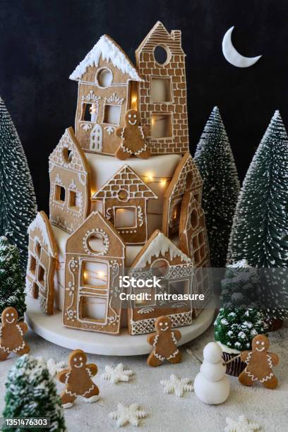 Image Of Homemade Tiered Christmas Cake Displayed On Cake Stand Cake Covered With Fondant Icing And Gingerbread House Biscuits Decorated With White Royal Icing Illuminated Fairy Lights Nighttime Christmas Village Scene Black Background And Crescent Stock Photo - Download Image Now