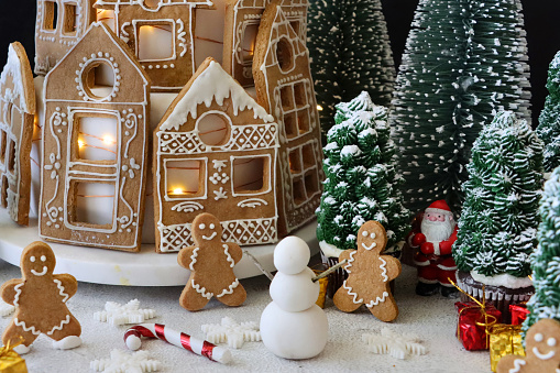 Stock photo showing close-up view of a night-time Christmas village scene designed Christmas cake. Homemade, tiered cake covered in fondant icing and gingerbread house-shaped cookies decorated with white royal icing displayed on cake stand Surrounded by cupcake Christmas trees.