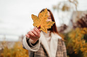 Young woman in coat and sweater holding in hand yellow maple leaf and cover face. Woman in park, forest. Fall season. Autumn blues, feeling down. Urban casual outfit. Lifestyle