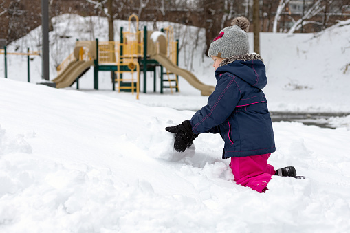 Small child making snowman, playing with snow near playground on snowy cold winter day. Girl wearing snowsuit outdoors. Fun outdoor activity for kids and family.