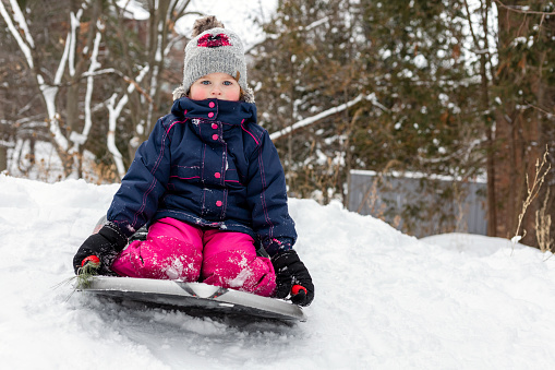 Little girl tobogganing downhill, having fun. Small happy child sledging in snow outdoors in winter. Little girl in hat and jacket sitting on sledge in cold weather.