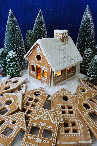 Stock photo showing close-up view of a snowy clearing in night-time, conifer forest scene. A homemade, gingerbread house decorated with white royal icing surrounded by Christmas tree cupcakes and gingerbread house biscuits on white, icing sugar snow.
