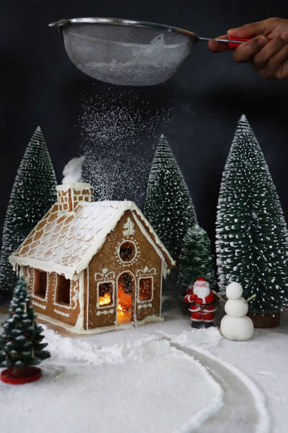 Image of unrecognisable person sifting powdered sugar over homemade, gingerbread house decorated with white royal icing displayed in snowy, night conifer forest scene illuminated by fairy lights, Santa Claus, snowman, icing sugar snow, black background Stock photo showing close-up view of a snowy clearing in night-time, conifer forest scene. A homemade, gingerbread house decorated with white royal icing is seen illuminated with fairy lights and surrounded by model fir trees on white, icing sugar snow against a black background. candy house stock pictures, royalty-free photos & images