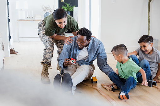 A female soldier sneaks up on her husband as he plays with toy trains with their two sons. The family smiles and laughs together.