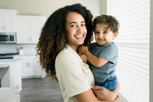 Beautiful photo of mother carrying son while standing near window Standing near a window in the living room, the beautiful mother and son smile for the camera. mom and baby eye contact stock pictures, royalty-free photos & images