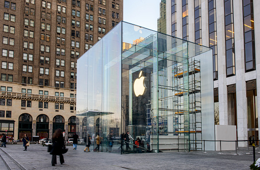 New York, United States - February 11, 2014: Glass building of the Apple Store with huge Apple Logo at 5th Avenue near Central Park. The store is designed as the exterior glass box above the underground display room. There customers waiting on the entrance and a few incidental people seen on the street.