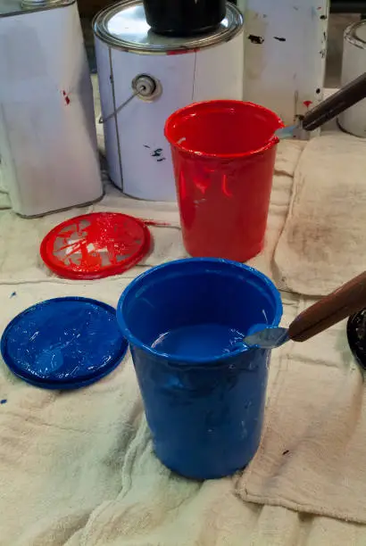 silkscreen ink with spatulas sitting in them. One cup full of red ink. One cup of blue ink. Cans of ink in the background