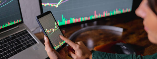 istock Horizontal banner or header close-up Unrecognizable woman holding smartphone touching screen to study graphics. Electronical devices and computers on background showing charts. 1351523037