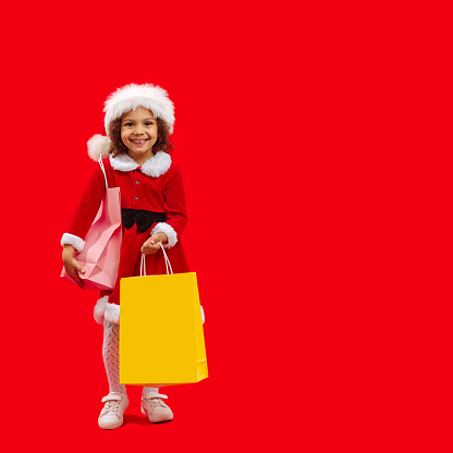 A little happy African American girl in a Santa hat and a carrier bag prepares gifts for friends before Christmas and New Year