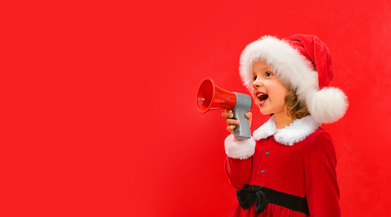 A child in a red Santa hat holds a megaphone in his hand and shouts. Concept for slogans for Christmas and New year, on a red background. Copy space.